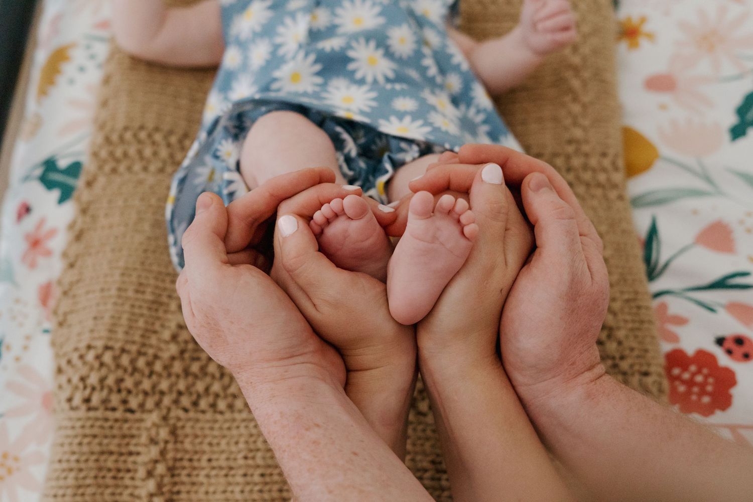 Two parents gently cradle a newborn babys tiny feet in their hands on a floral blanket.