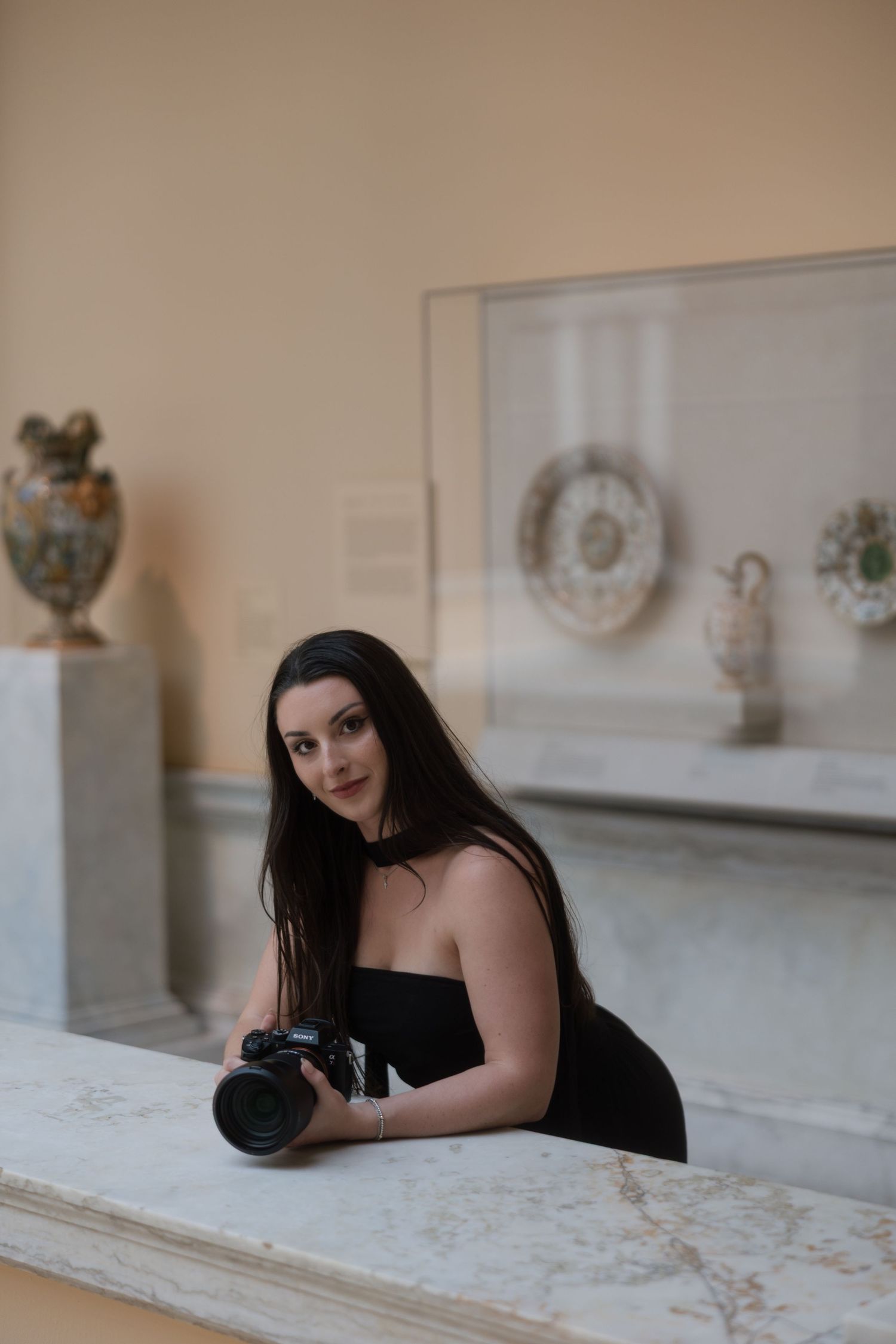 Young woman in black dress holding a DSLR camera, leaning on marble ledge in an art museum with antique ceramics display.