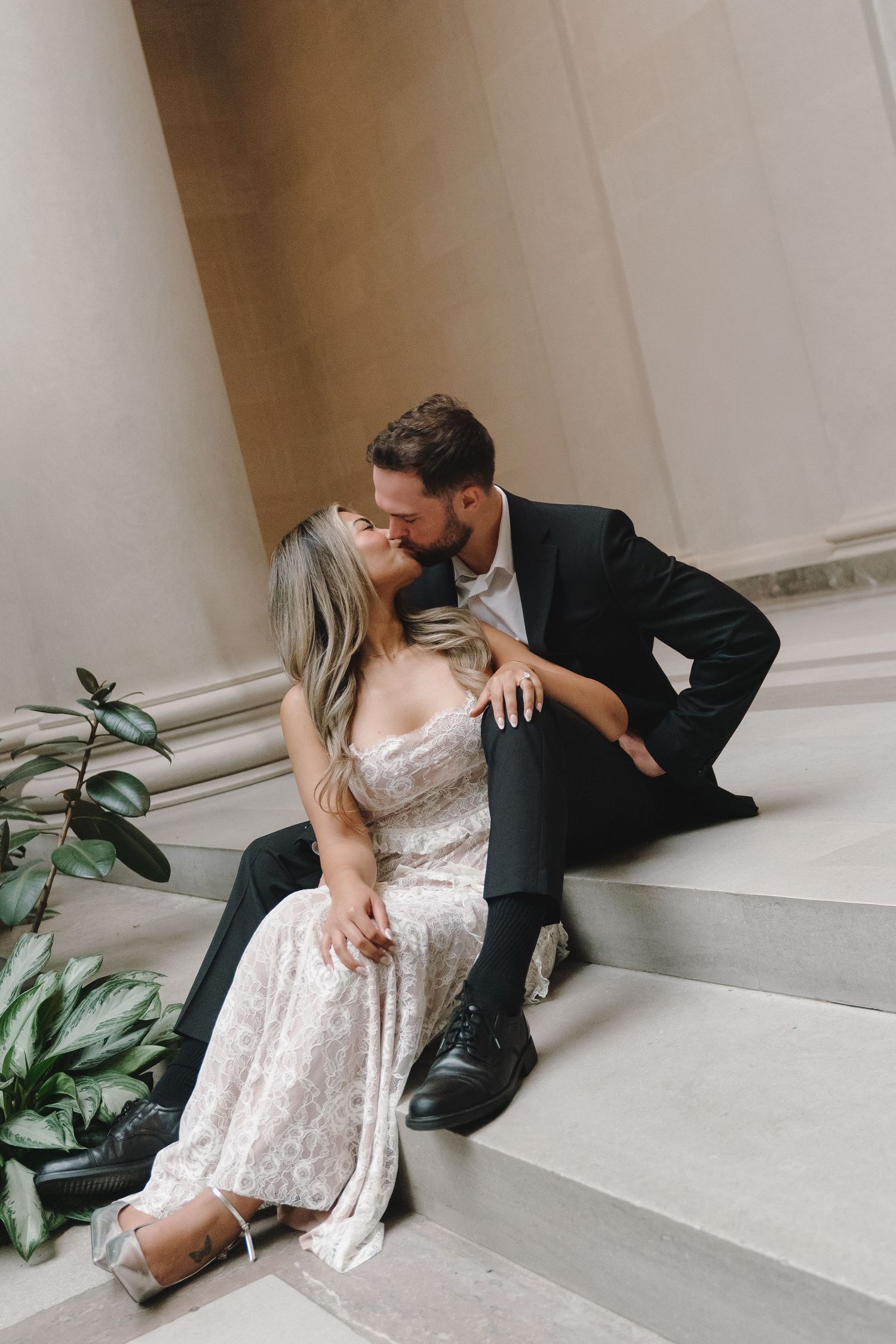 Couple sharing a romantic kiss while sitting on marble steps, woman in lace gown and man in black suit.