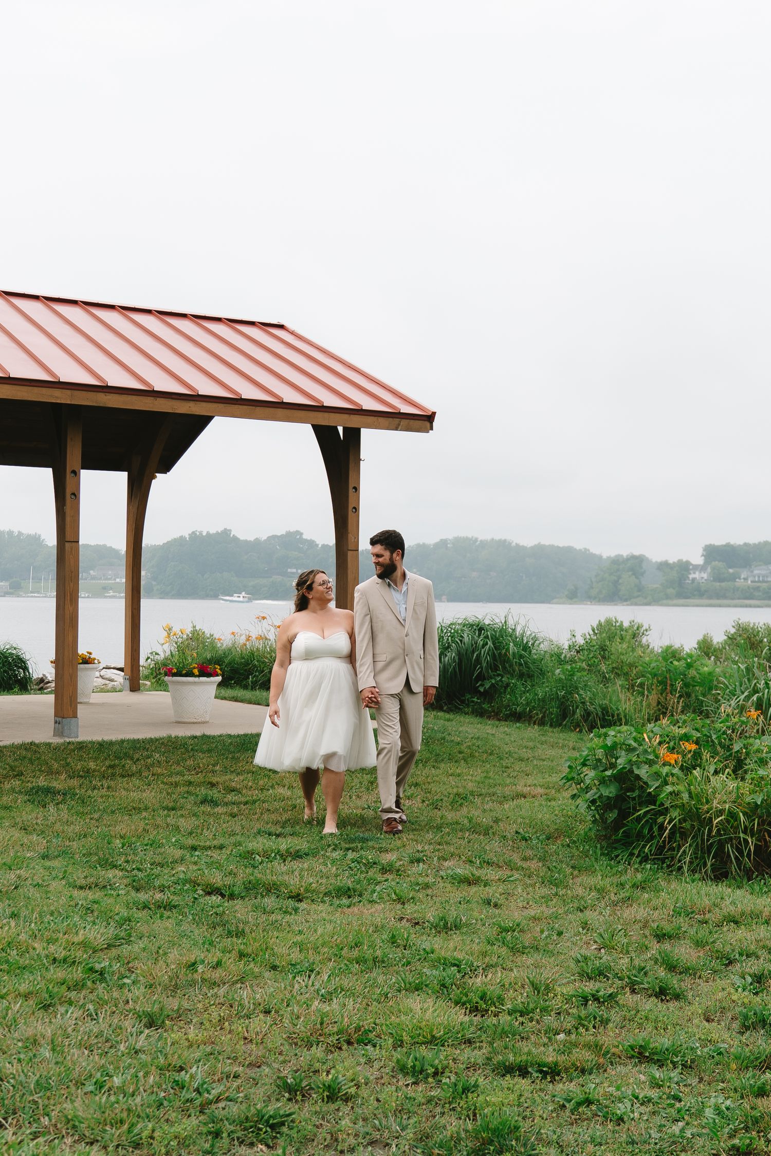 Bride and groom walk hand in hand on green lawn near a wooden pavilion with red roof by a lake.