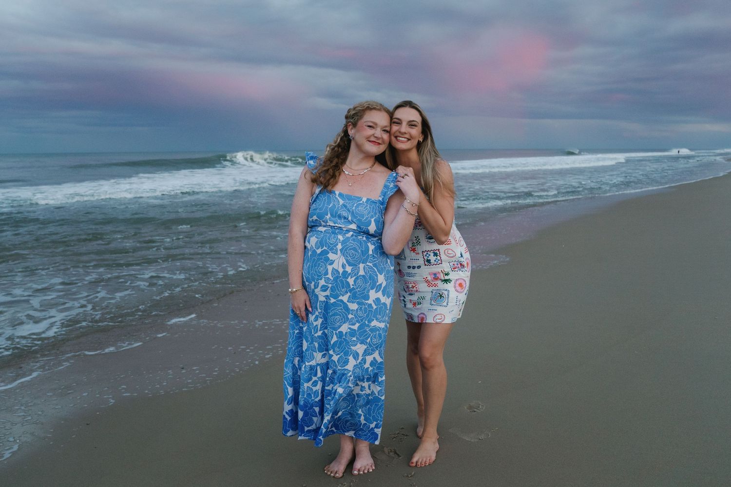 Two women smiling together on a beach at sunset with colorful pink and blue sky behind them.