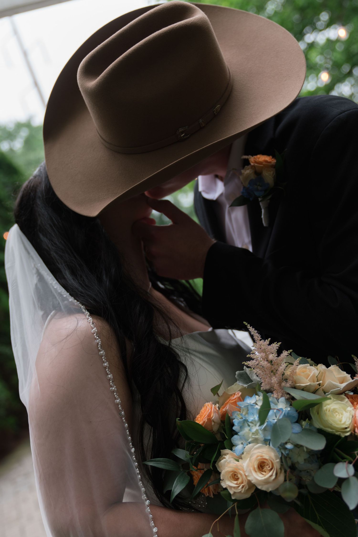 Bride and groom sharing a kiss under a cowboy hat with a pastel wedding bouquet outdoors.