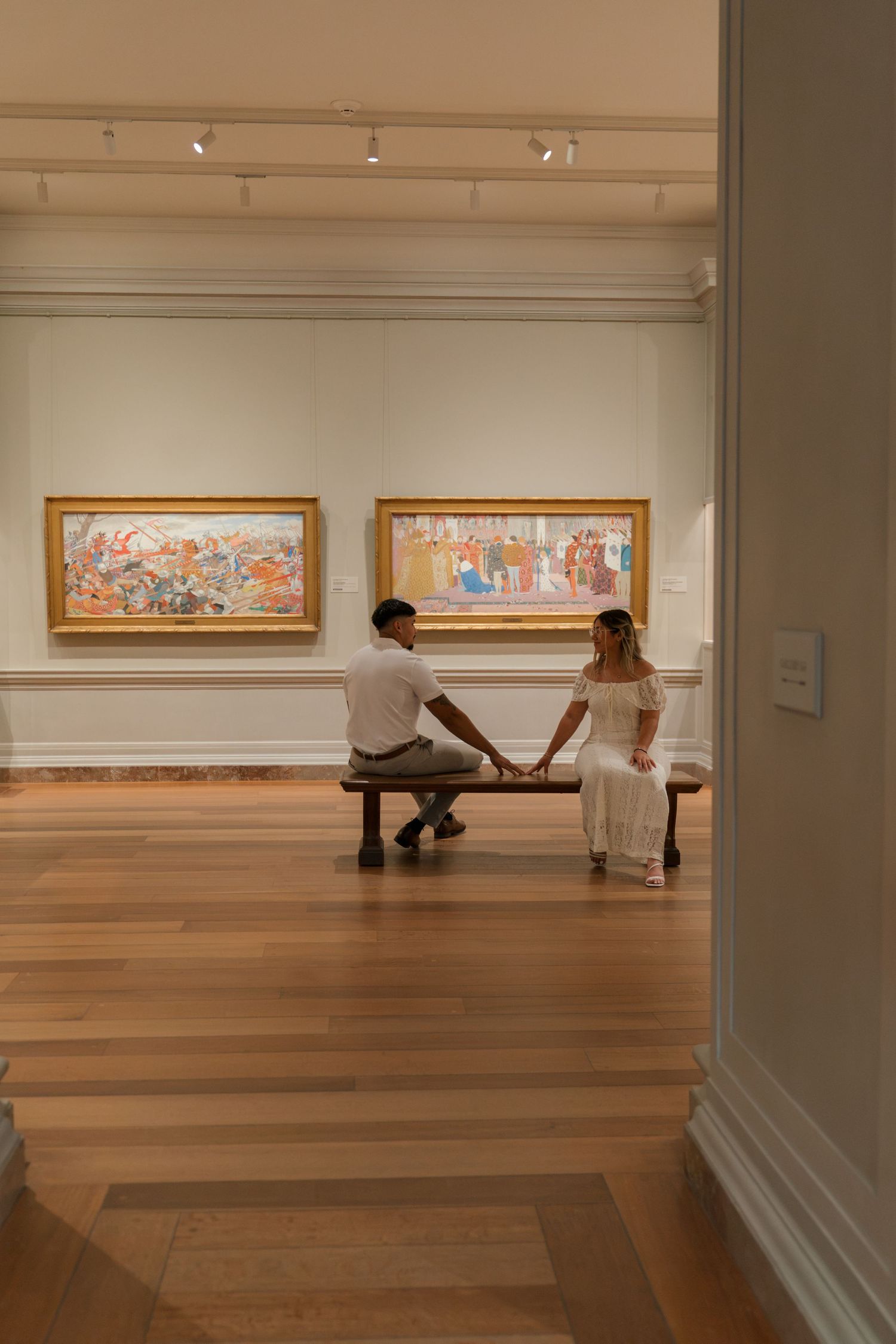 Couple holding hands on a bench in an elegant art museum gallery with colorful framed paintings on white walls.