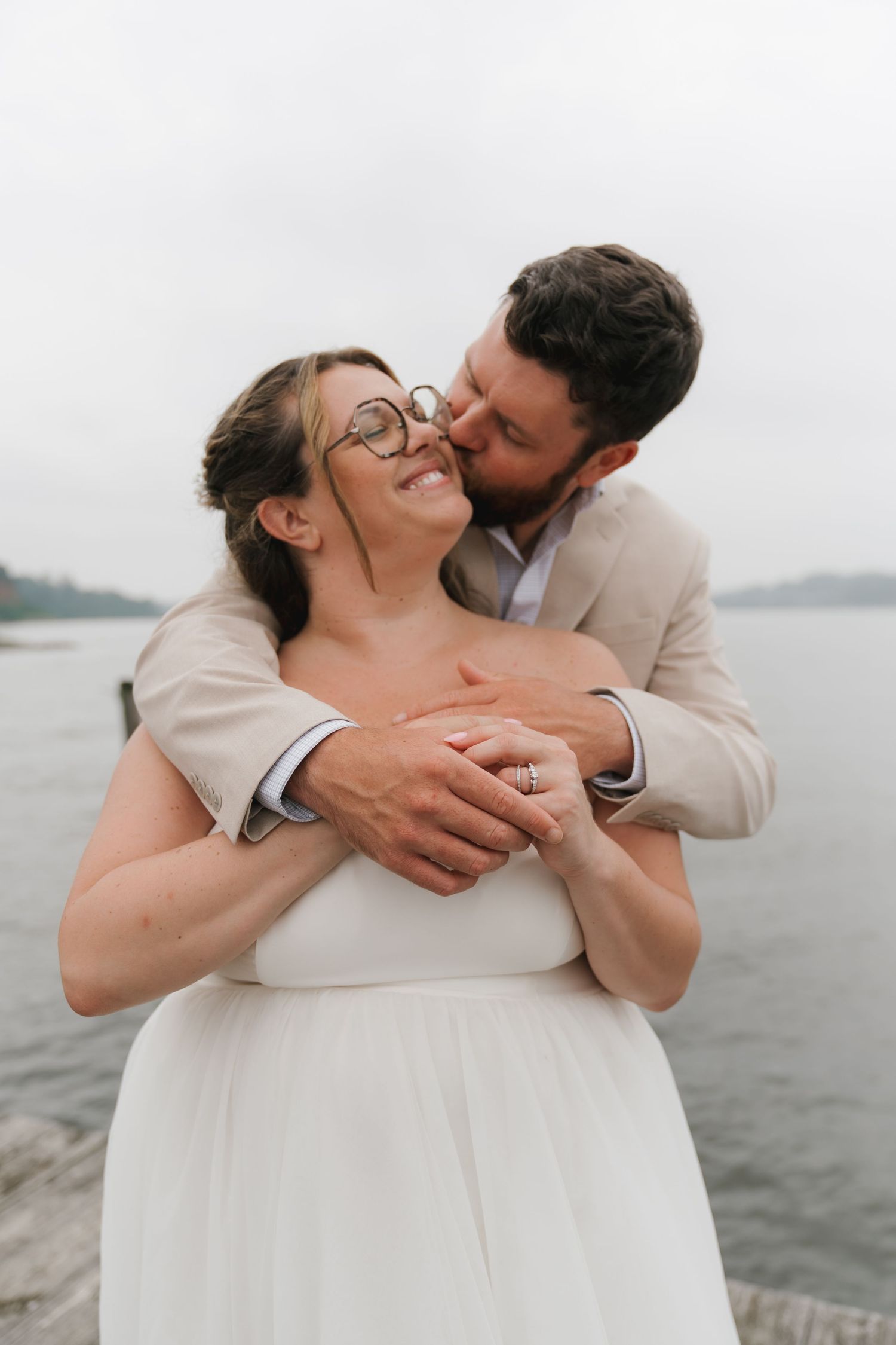 Happy bride wearing glasses smiles as groom in beige suit hugs her from behind near a serene lake.