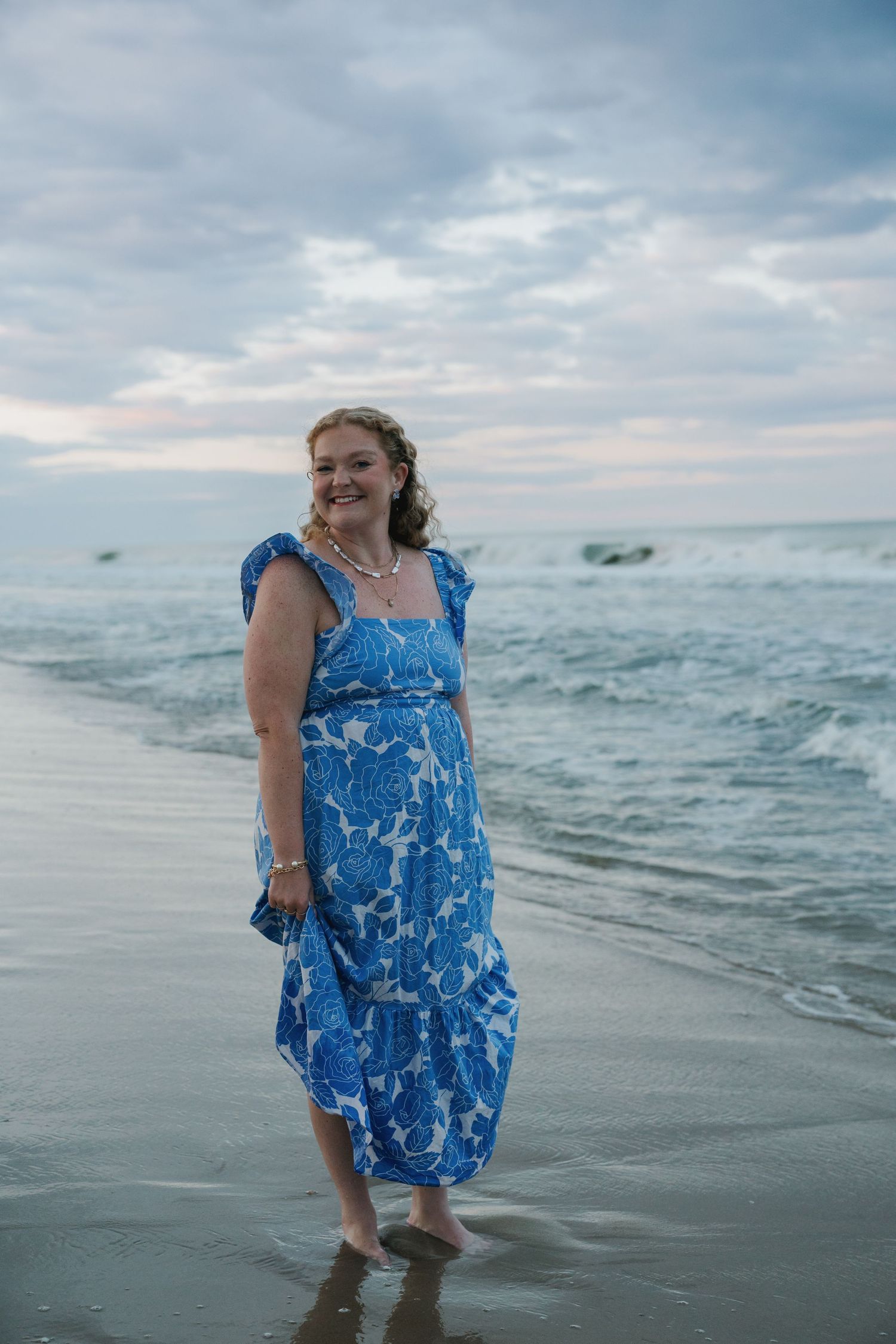 Woman in a blue floral dress standing barefoot in ocean waves on a beach under a dramatic cloudy sky.