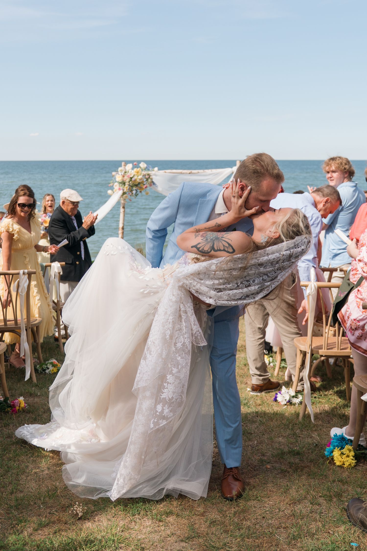 Groom dips bride for a romantic kiss at outdoor beach wedding ceremony with guests celebrating.