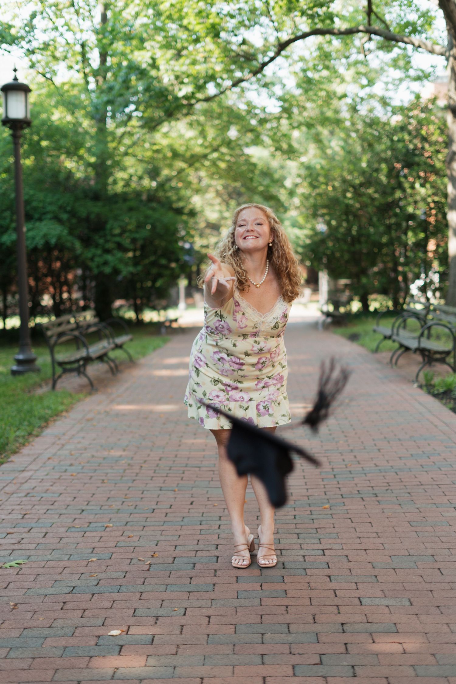 Smiling woman in floral dress tossing graduation cap on brick pathway lined with benches and green trees.