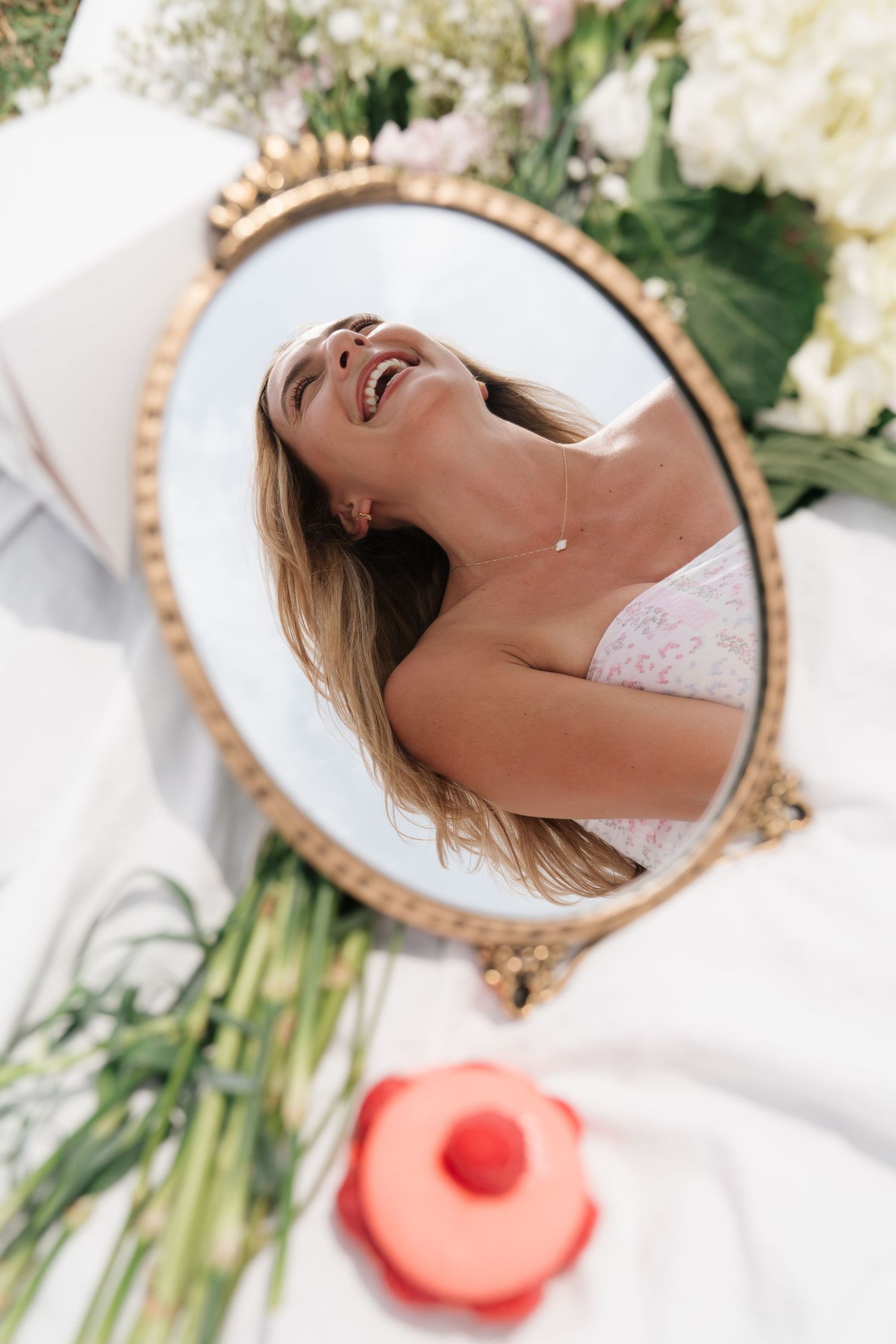 Woman laughing reflected in vintage gold oval mirror surrounded by white flowers and greenery outdoors.
