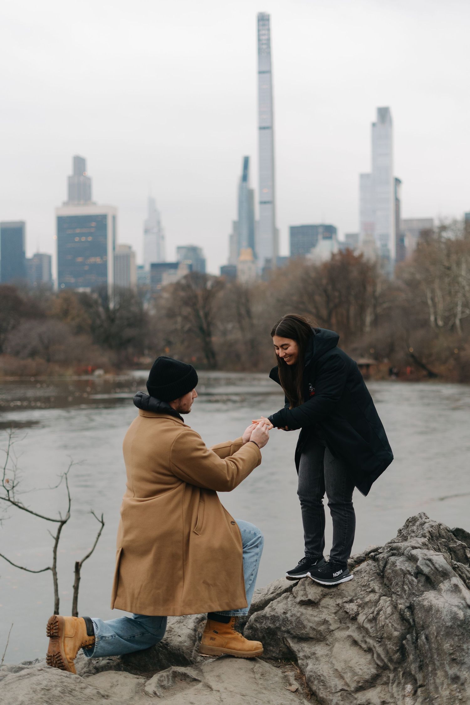Man proposing on one knee to woman on rocky lakeside with NYC skyline backdrop.