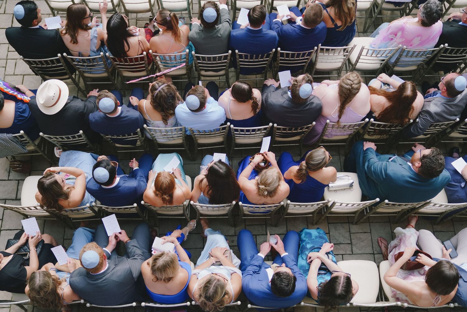 Aerial view of wedding guests seated in rows of chairs at an outdoor ceremony, holding programs.