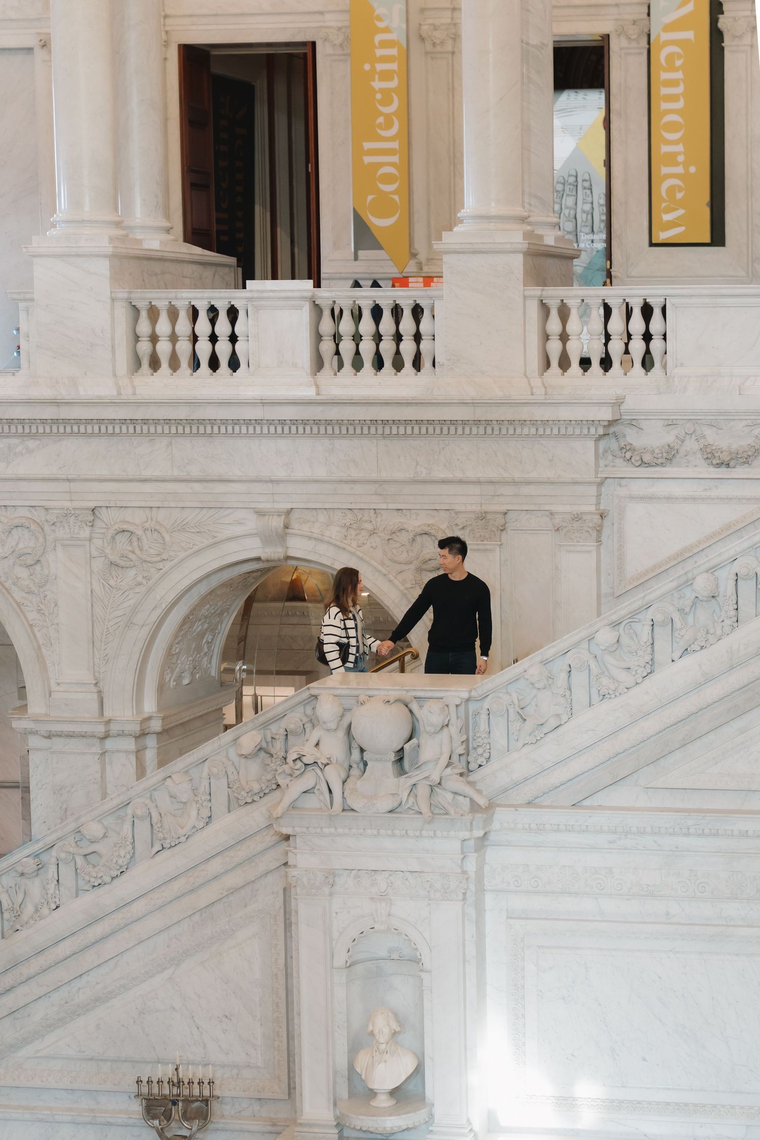 Couple holding hands on ornate marble staircase inside a grand neoclassical building.