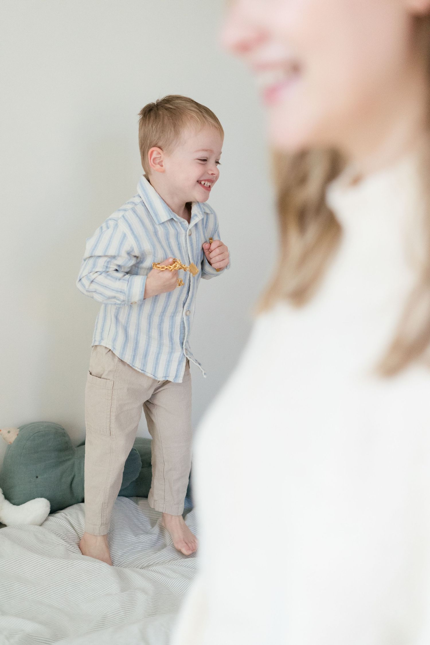 Joyful toddler boy in striped shirt jumps on bed laughing during family lifestyle photo session.