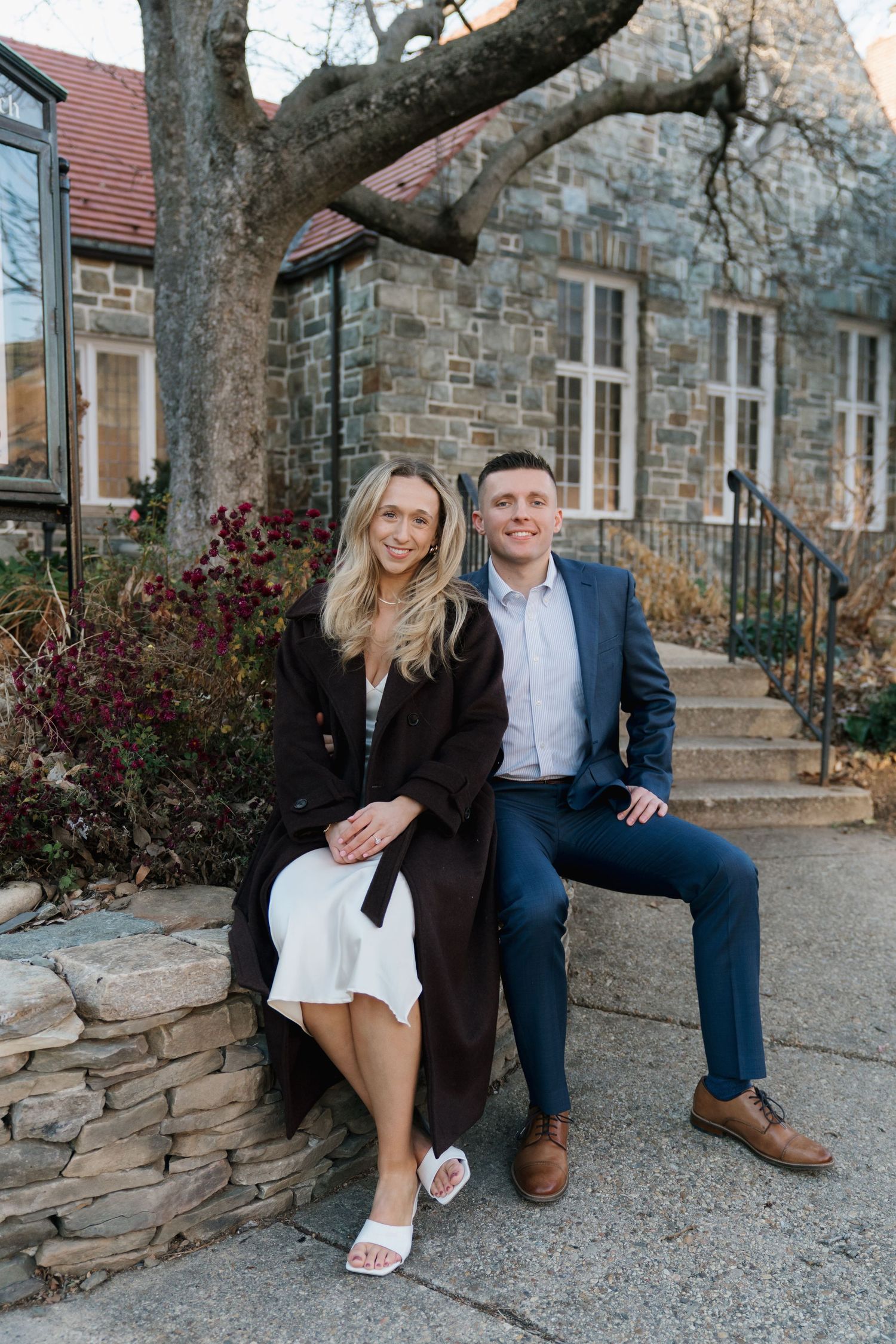Engaged couple sitting together on a stone wall outside a historic stone building.