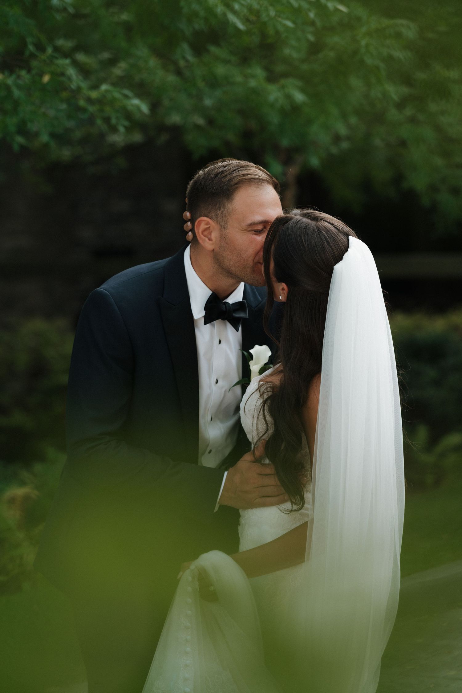 Groom in navy tuxedo kissing bride in white veil surrounded by lush green garden foliage.