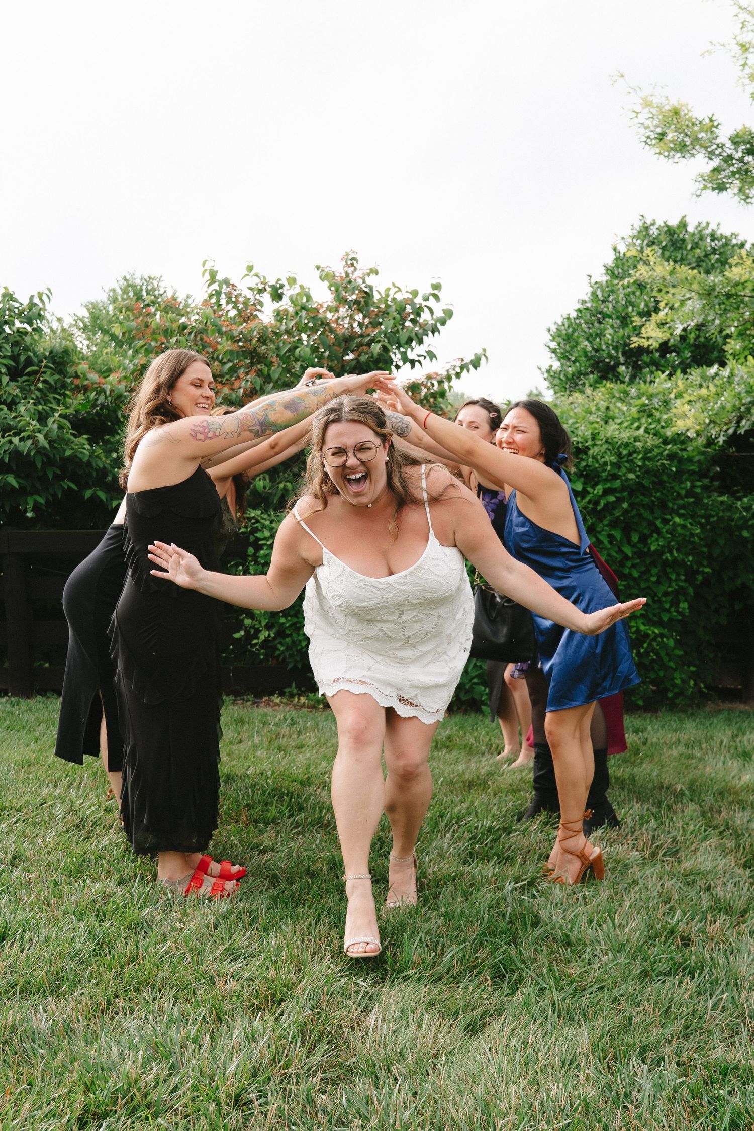 Bride laughing as bridesmaids playfully dance around her on a green lawn at a wedding reception.