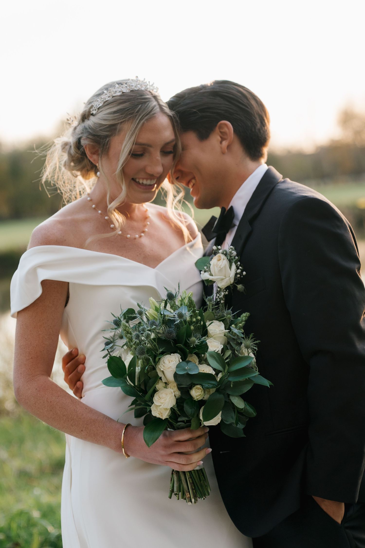 Smiling bride in off-shoulder gown and groom in tuxedo sharing a tender moment with white bouquet.