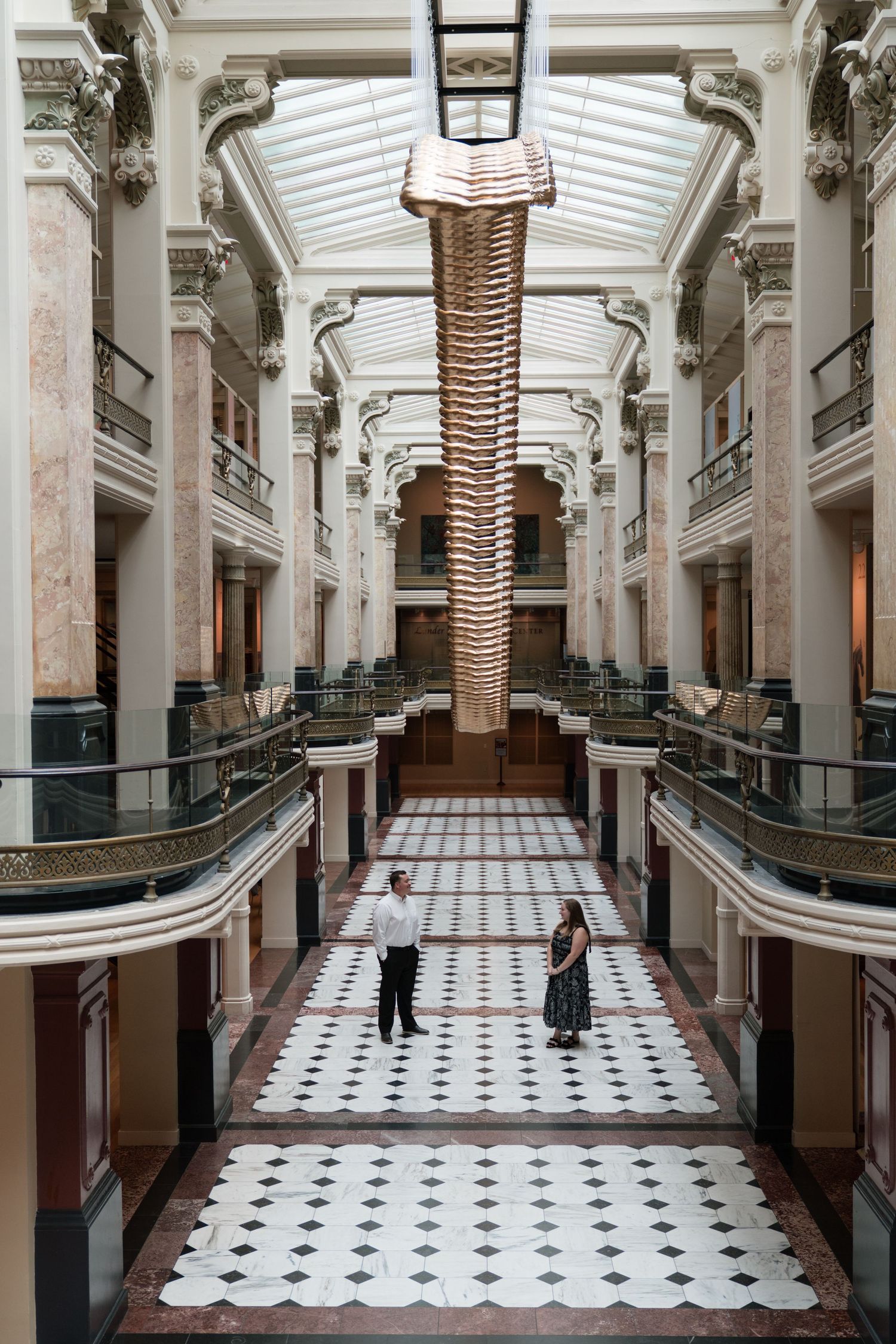 Two people stand beneath a large vertebrae sculpture in an ornate multi-story atrium with marble floors.