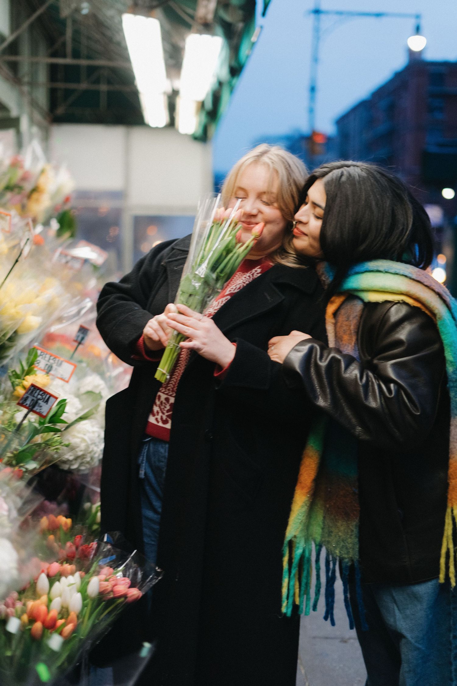 Two women embracing at an outdoor flower market, one holding a bouquet of white tulips.