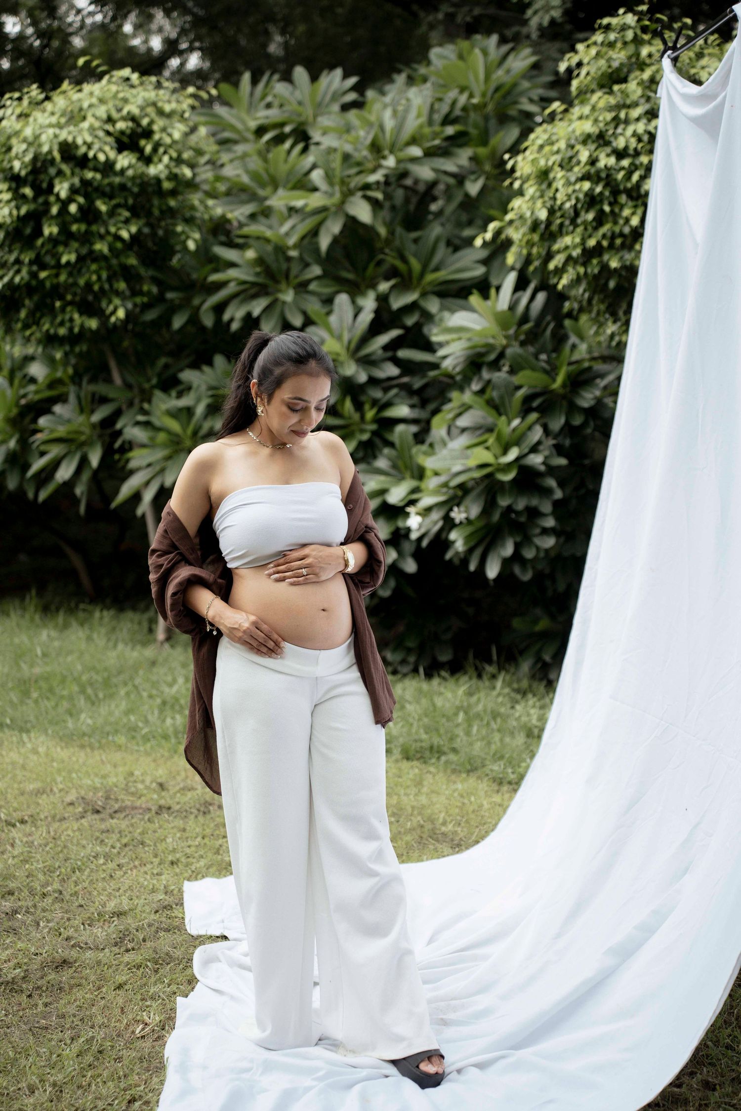 Expectant mother in white outfit and brown cardigan poses outdoors against lush greenery and flowing white fabric backdrop.