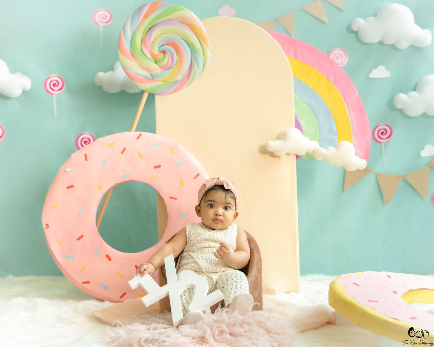 Baby girl in white dress sits by giant donut prop with rainbow and candy decorations in a colorful studio setting.