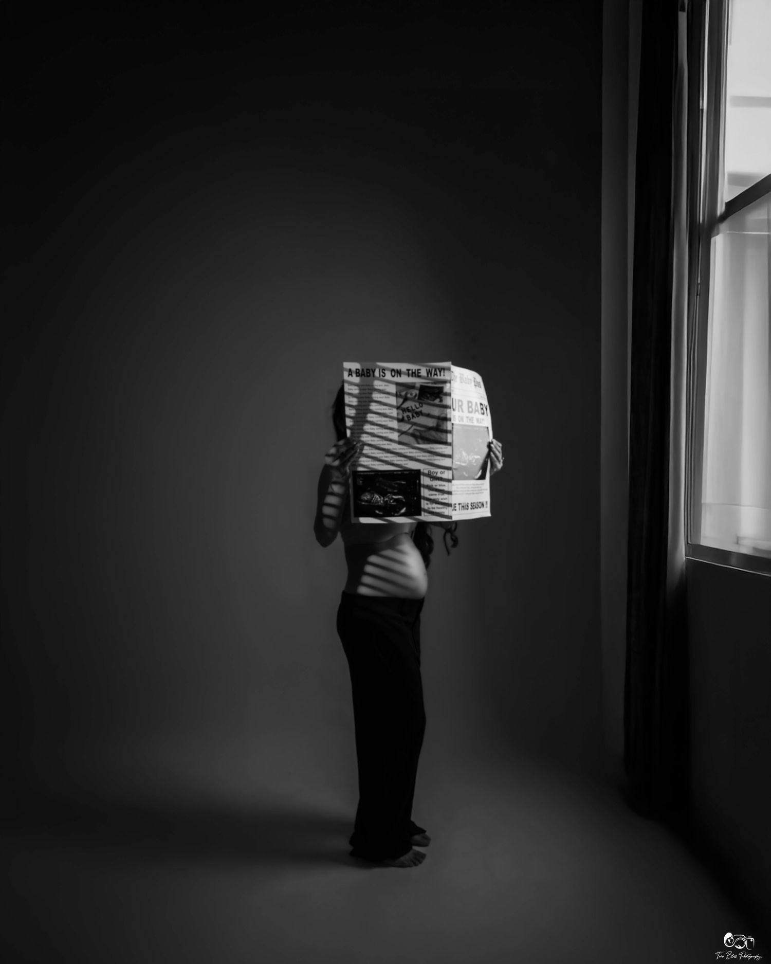 Black and white image of figure holding an accordion against a dark wall near a window.