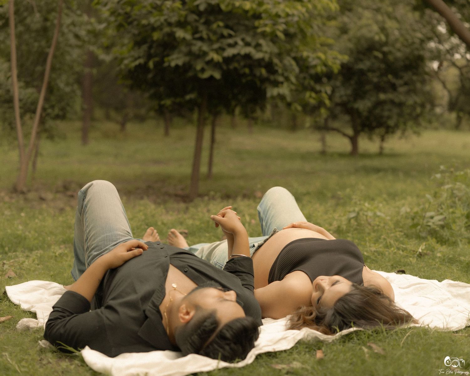 Two people lying on a white blanket in a lush green park, relaxing outdoors on a sunny day.