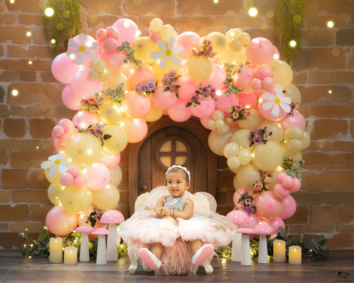 Baby girl sits smiling in front of a floral balloon arch with pink and yellow balloons and candles.