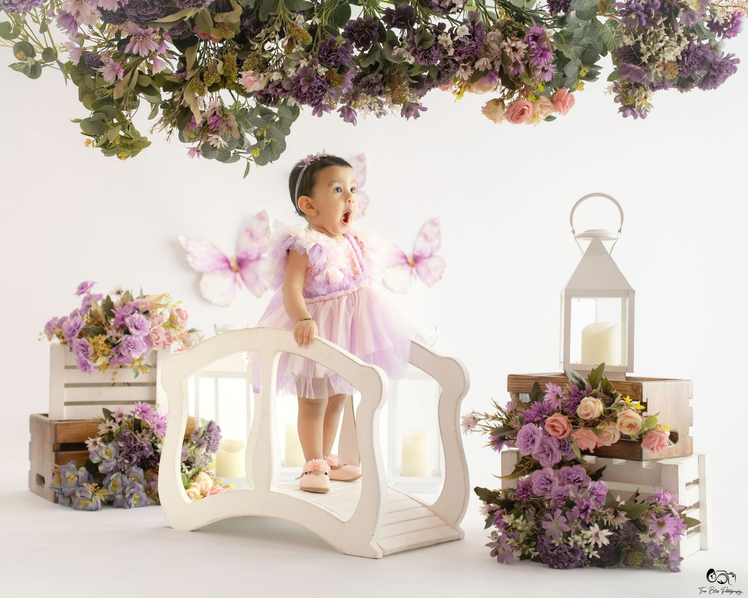 A baby girl in a pink fairy dress with butterfly wings stands on a white bridge surrounded by colorful flowers.