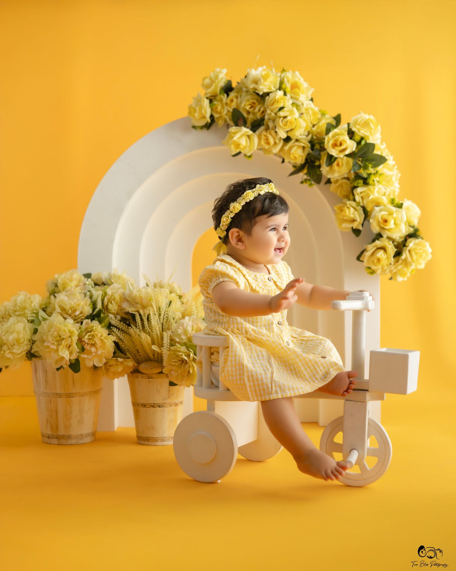 Baby in yellow dress sleeping among floral arrangements and white decor against bright yellow backdrop.