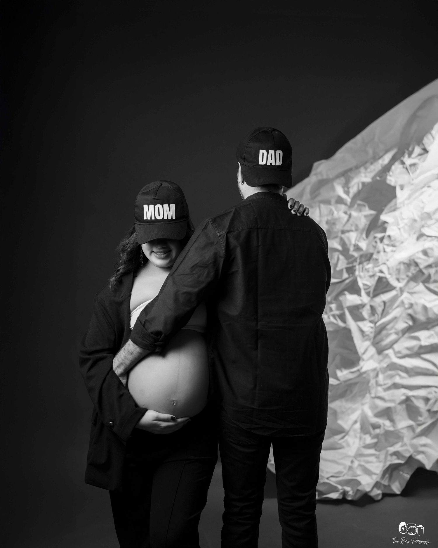 Black and white maternity photo of couple wearing MOM and DAD caps, pregnant woman cradling baby bump in studio.