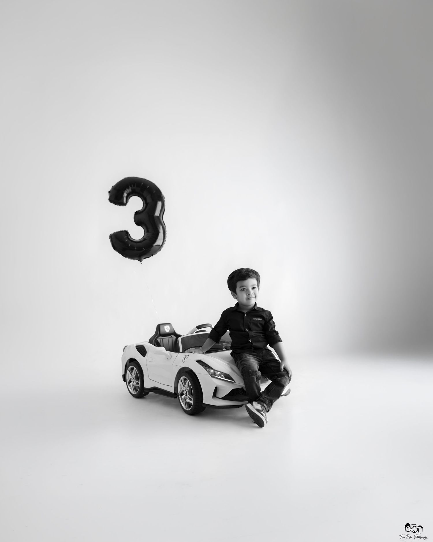 Black and white photo of a young boy sitting on a toy car beside a number 3 balloon, celebrating his 3rd birthday.