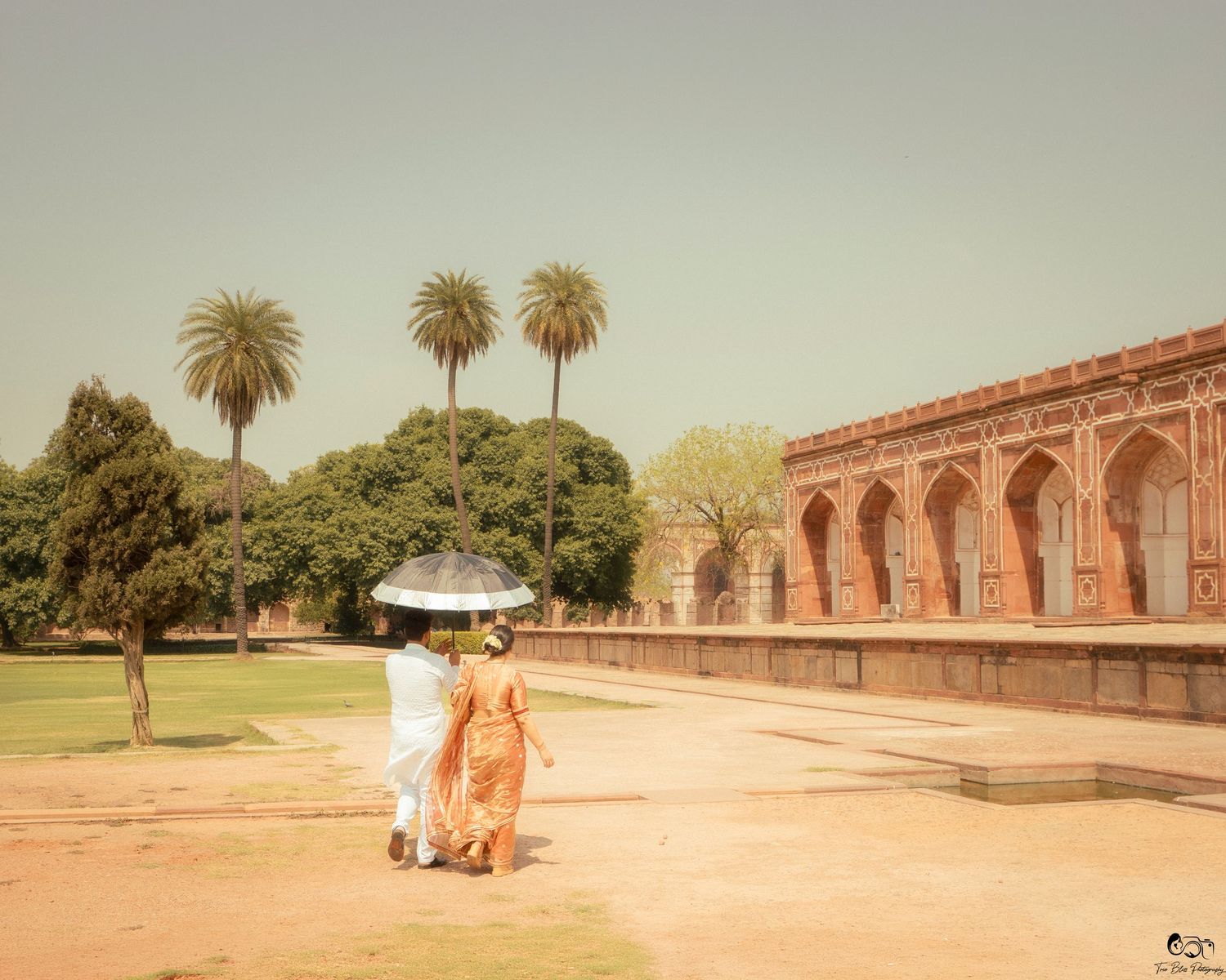 Couple walking with umbrella at Humayun's Tomb, Delhi, India, surrounded by palm trees and Mughal architecture.