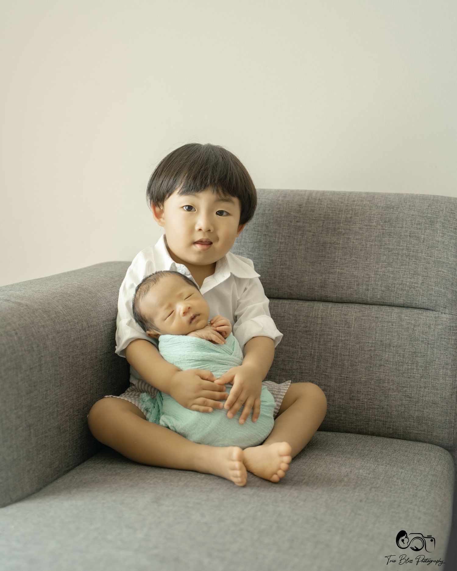 Young boy sitting cross-legged on gray sofa gently holding sleeping newborn baby wrapped in mint green swaddle.