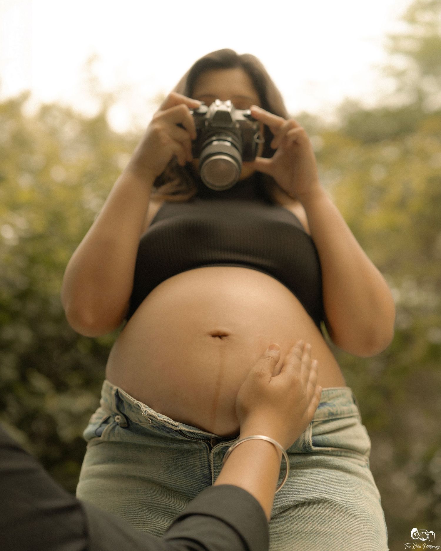 Pregnant woman in black crop top and jeans holding a camera, with a hand gently touching her baby bump outdoors.