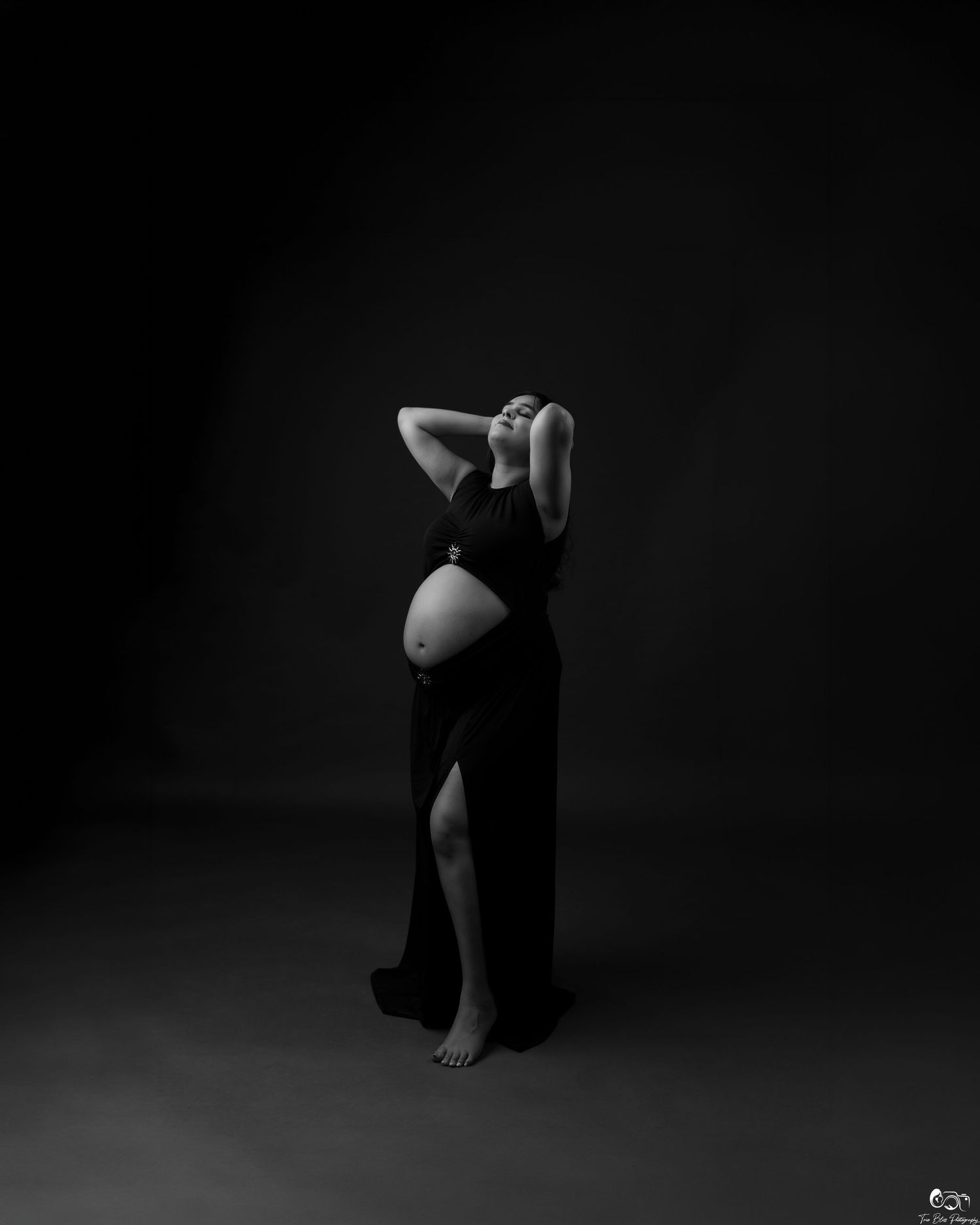 Black and white maternity photo of a pregnant woman in a black dress posing elegantly against a dark studio background.