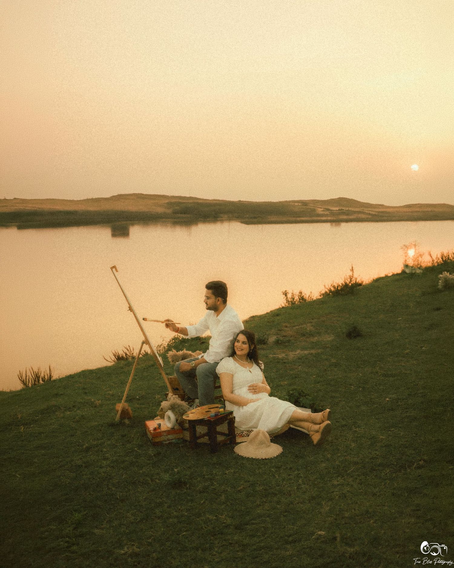 Couple in white outfits enjoying a romantic sunset picnic by a serene lake, with a easel and basket on green grass.