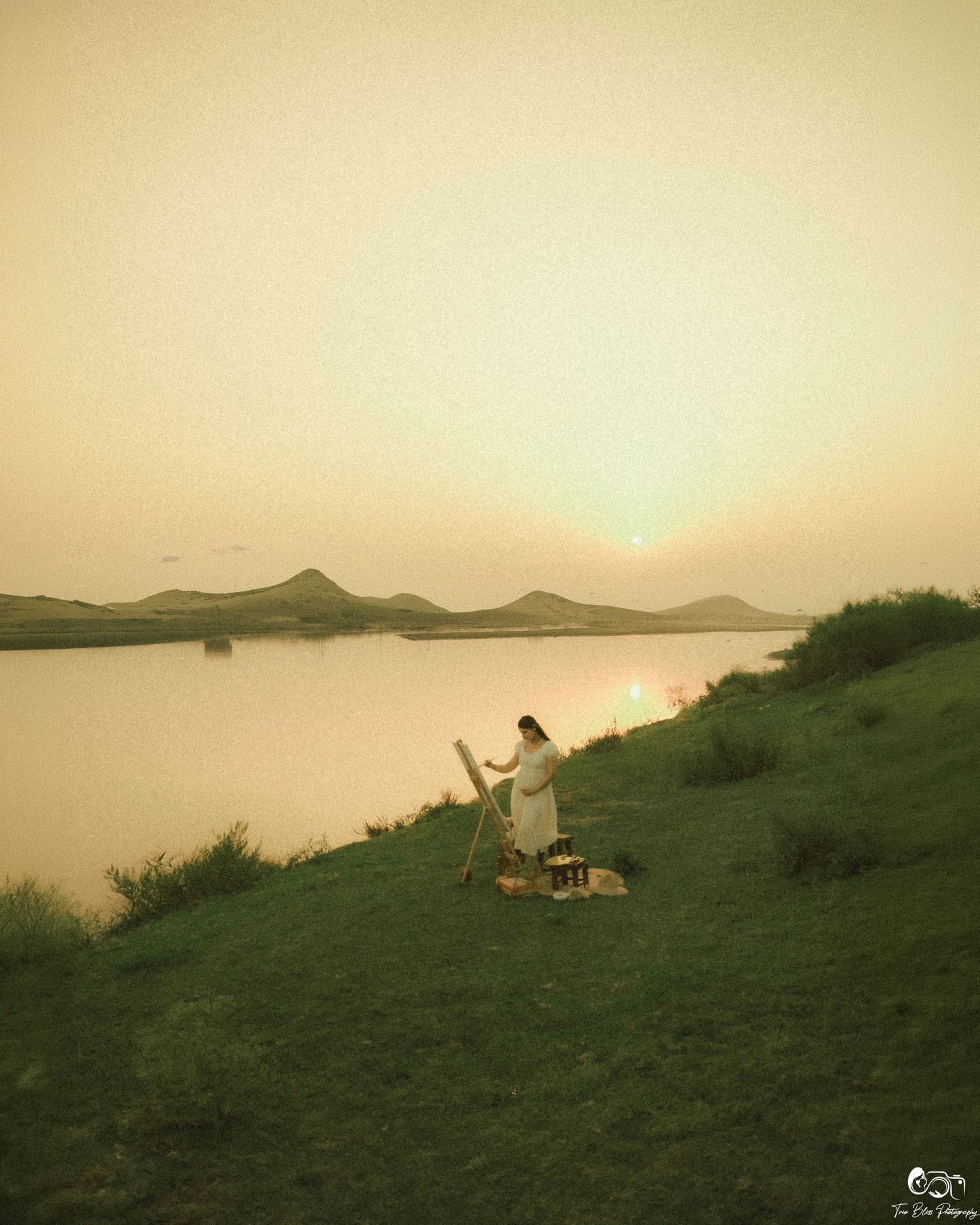 Artist painting on canvas at sunset by a calm river with mountains in the background on a grassy hillside.