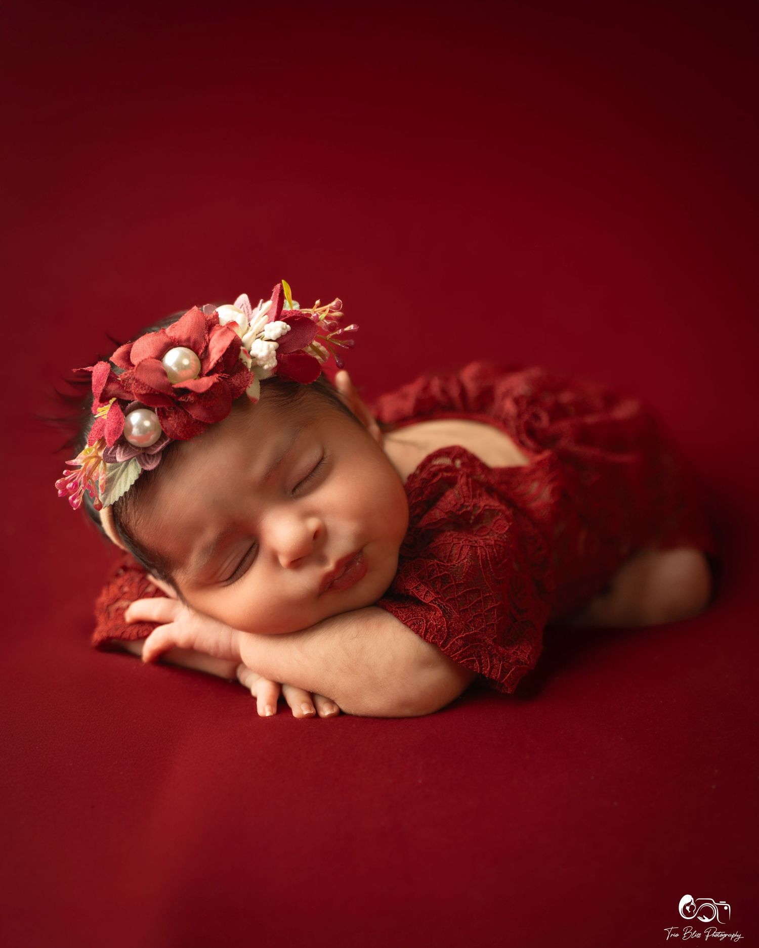 Sleeping infant in red lace outfit with pink and white floral crown against deep burgundy background.