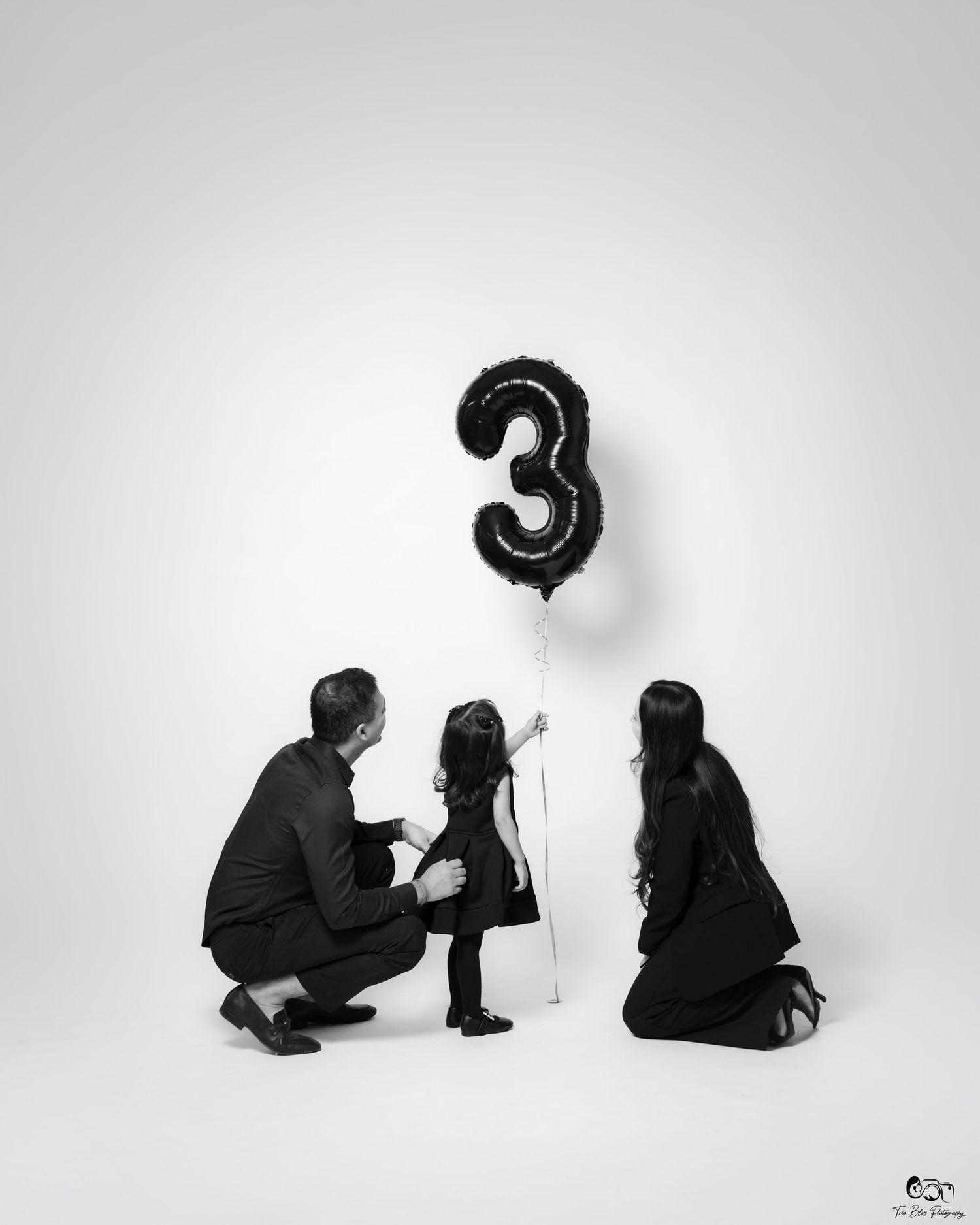 Black and white photo of parents crouching with toddler holding a number 3 balloon, celebrating a third birthday.