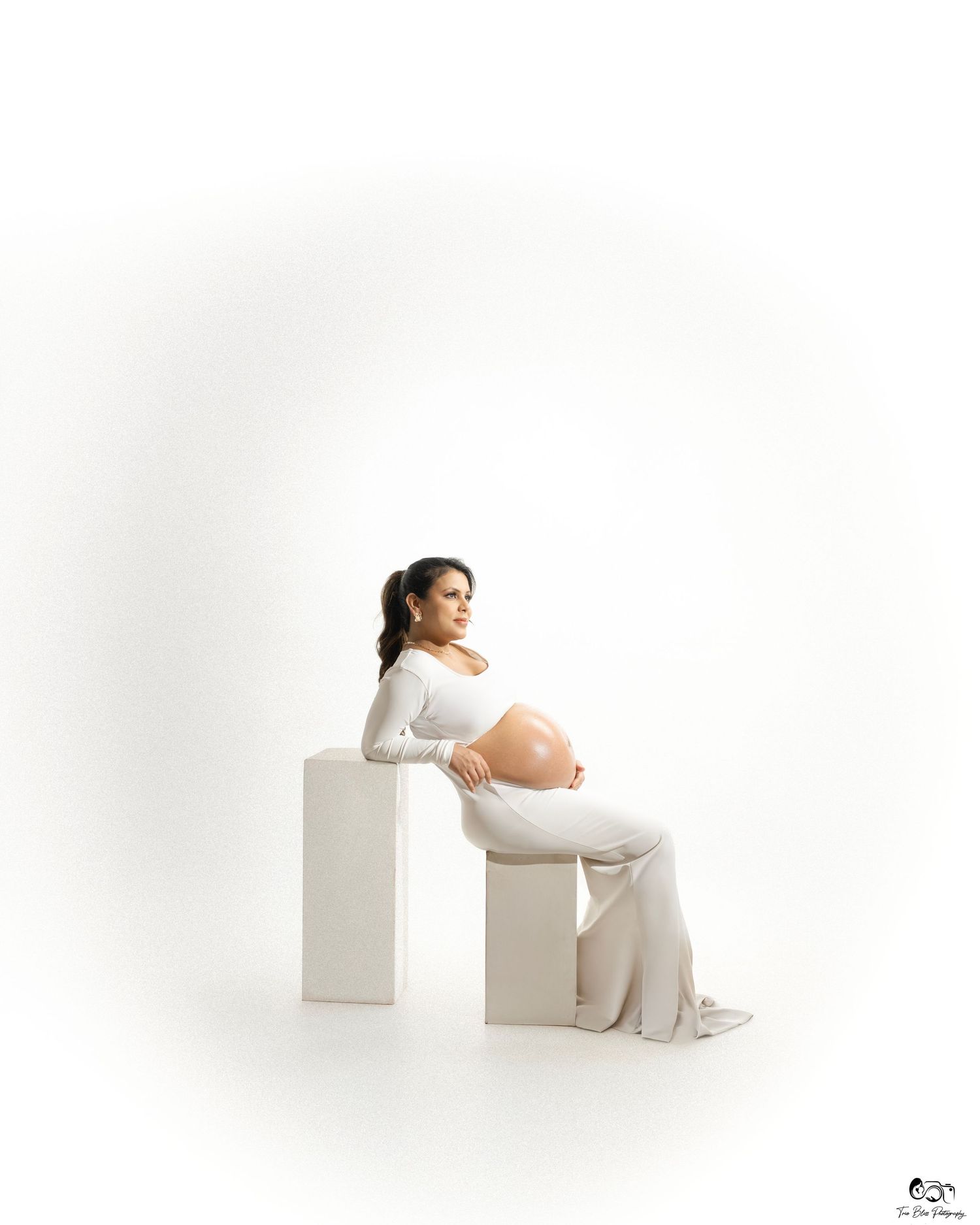 Pregnant woman in white outfit poses elegantly on white blocks during a professional maternity photo shoot.