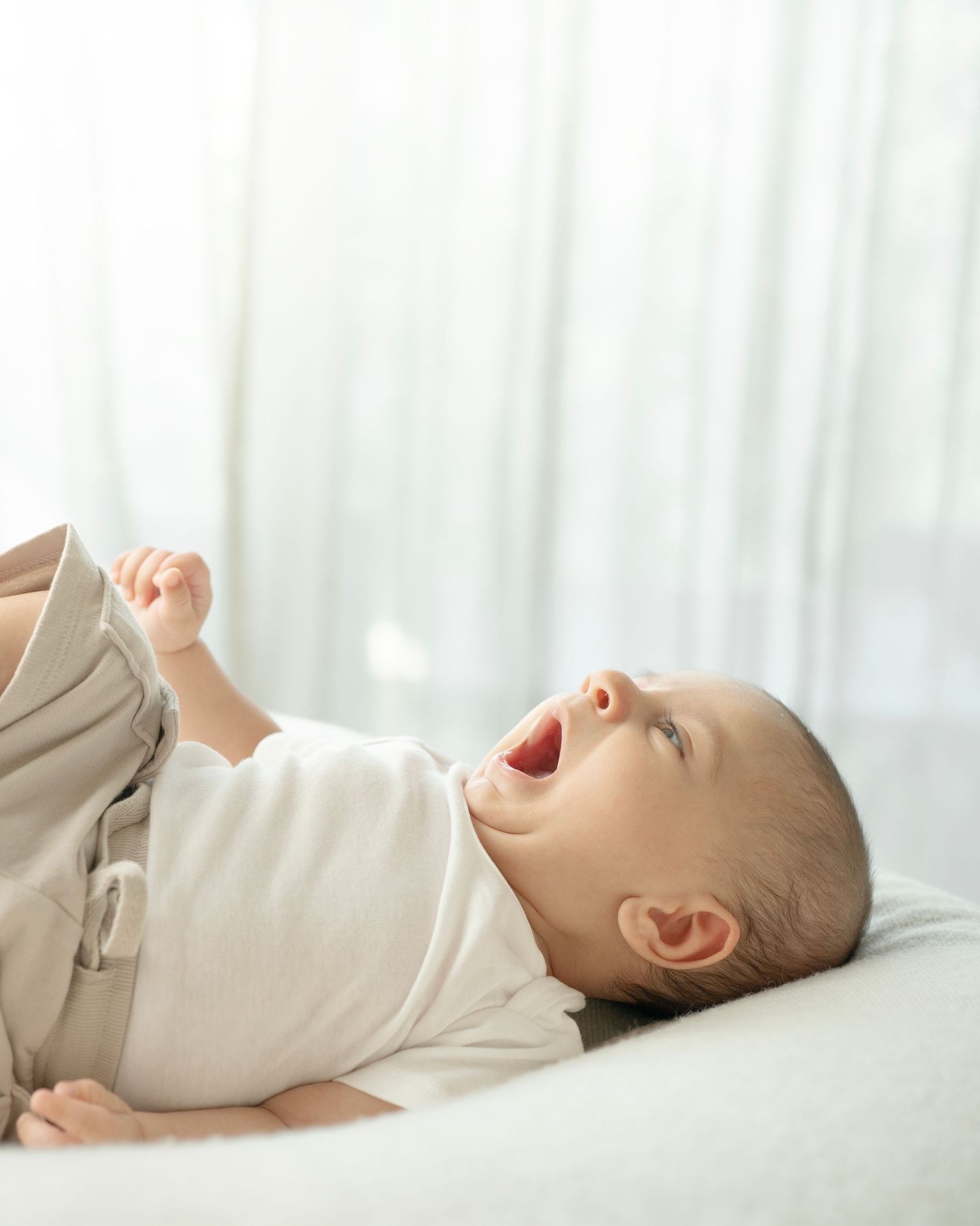 A happy baby lying on its back, mouth open in a yawn, wearing a white onesie and beige pants.
