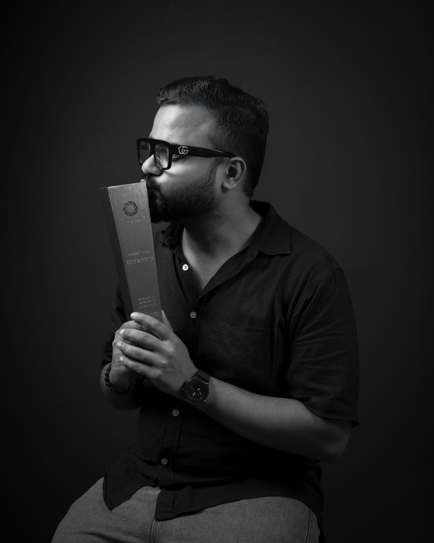 Black and white portrait of someone in glasses holding and kissing a book or award against a dark background.