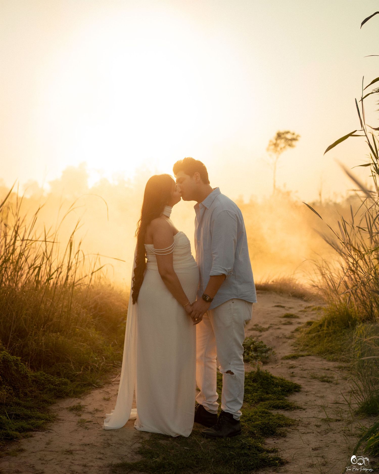 A couple shares a romantic moment at golden hour sunset, woman in white maternity gown holding hands with man in white outfit.