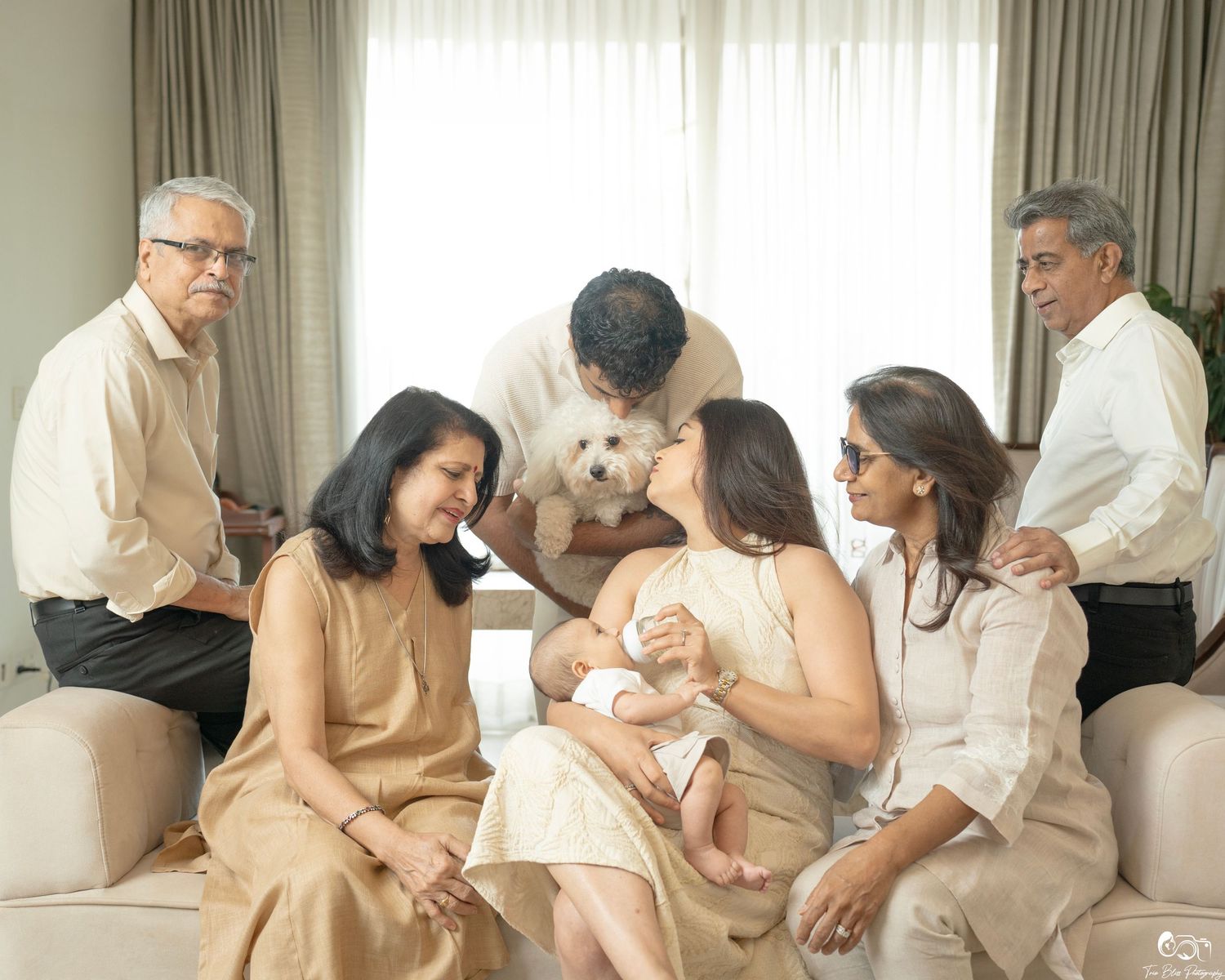 Multi-generational Indian family gathered around a mother nursing a newborn baby on a sofa in a bright living room.