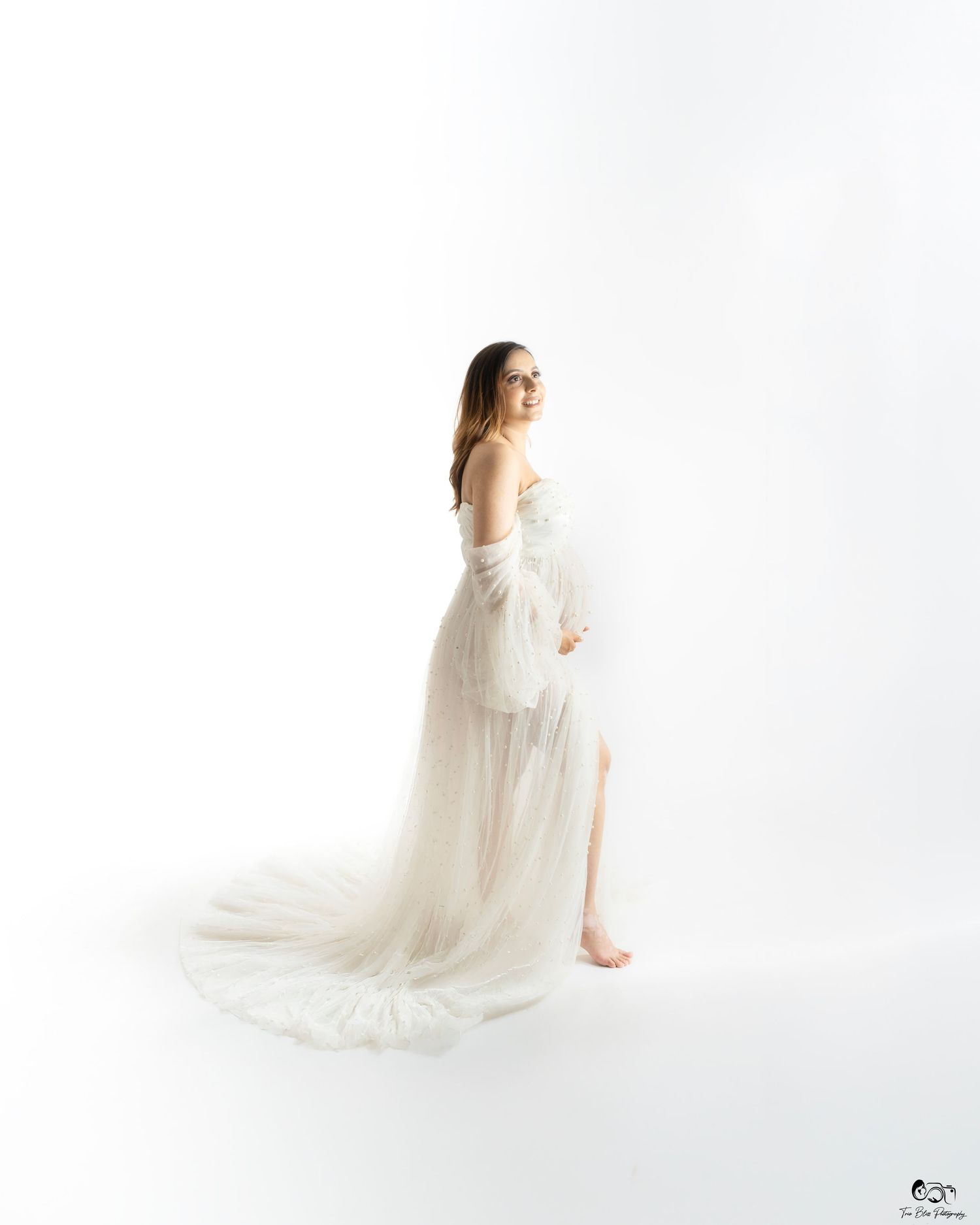 Pregnant woman in elegant white tulle gown with flowing train poses gracefully in a bright studio setting.