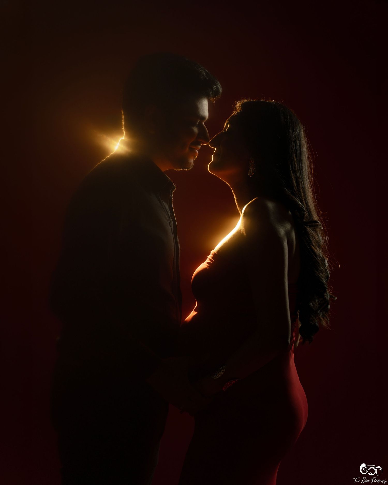 Romantic couple silhouetted against warm golden backlight in an intimate pose against a dark red background.