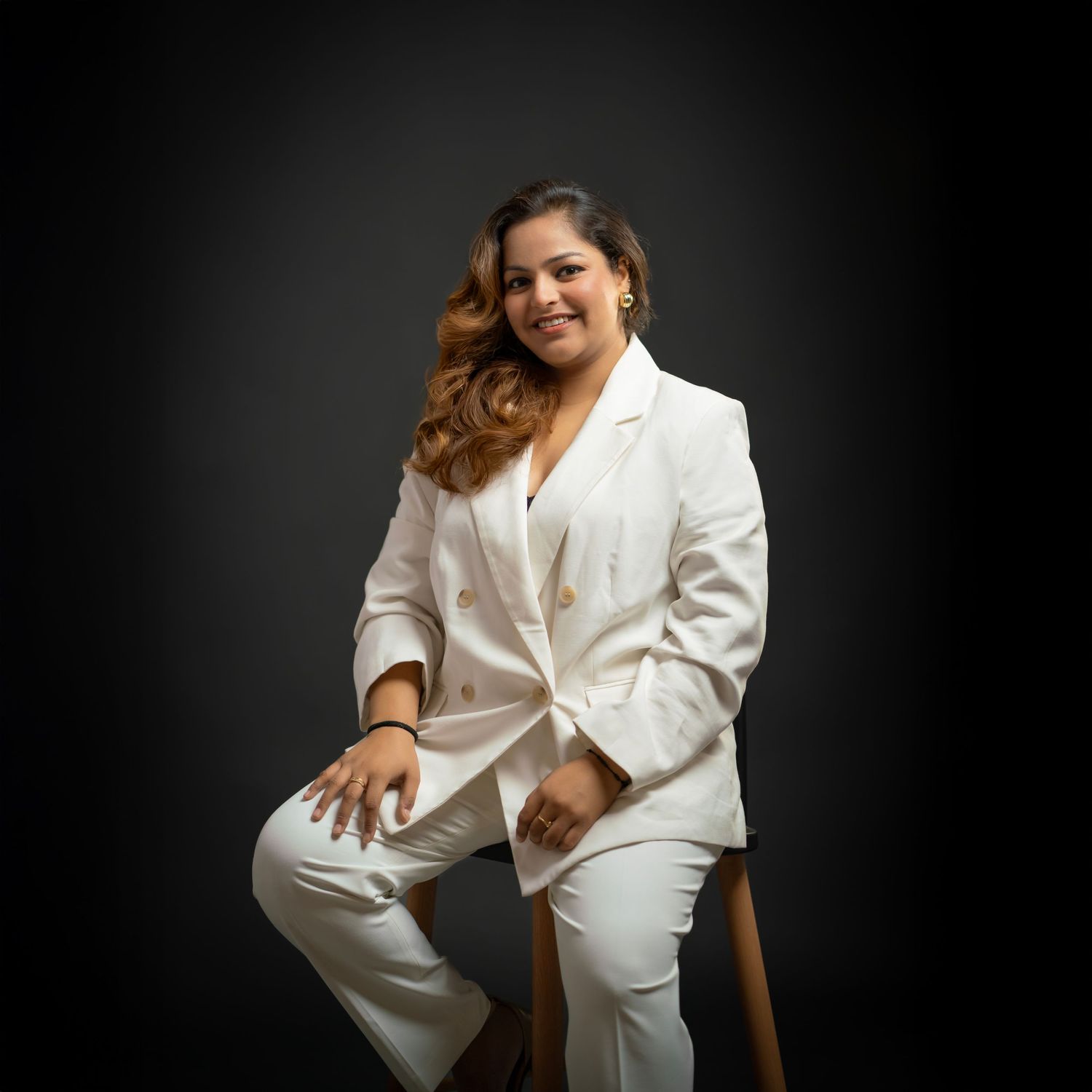 Professional portrait of someone in an elegant white pantsuit seated on a stool against a dark background.