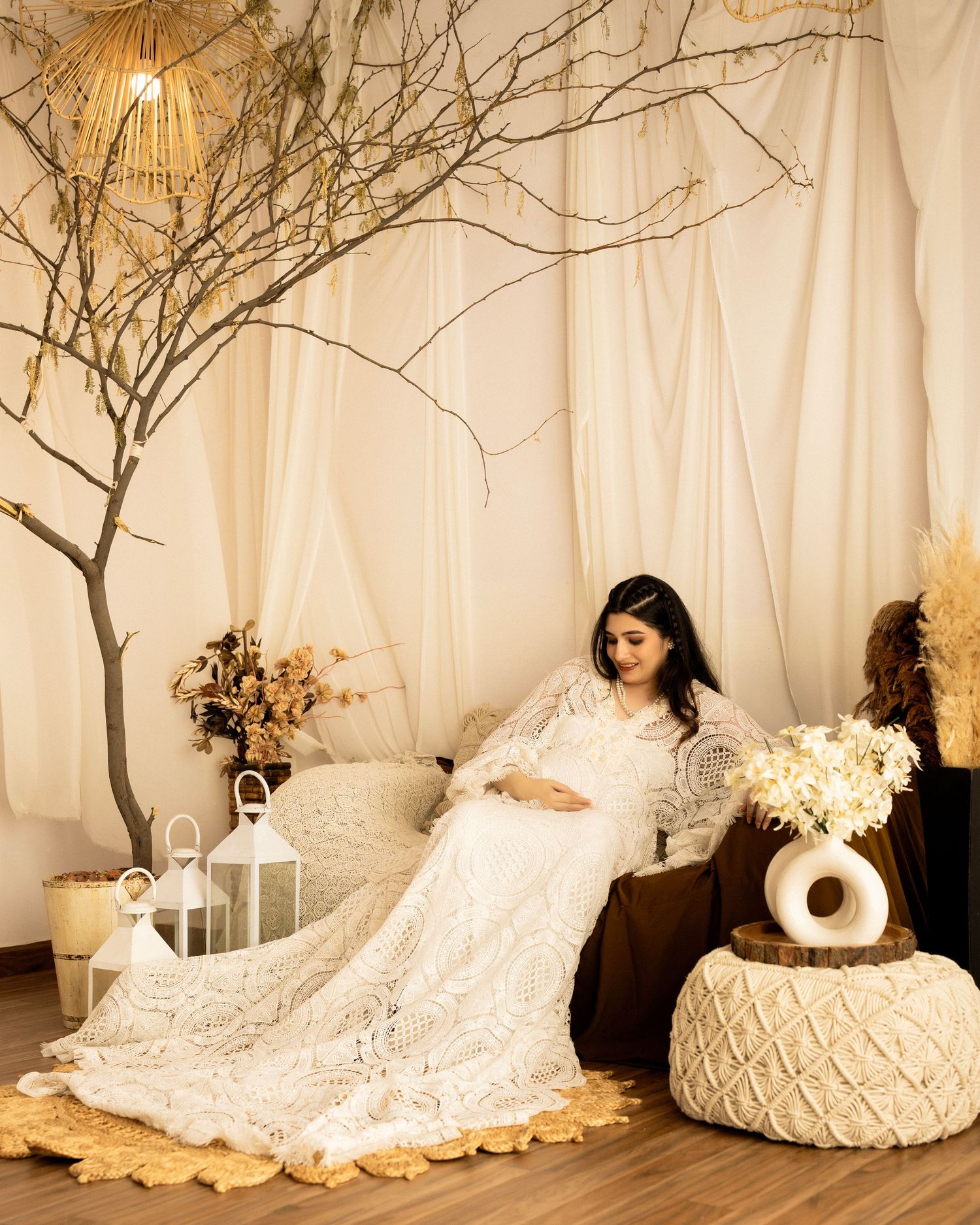 Ethereal maternity photo in white lace dress with decorative branches and lanterns in a dreamy studio setting.