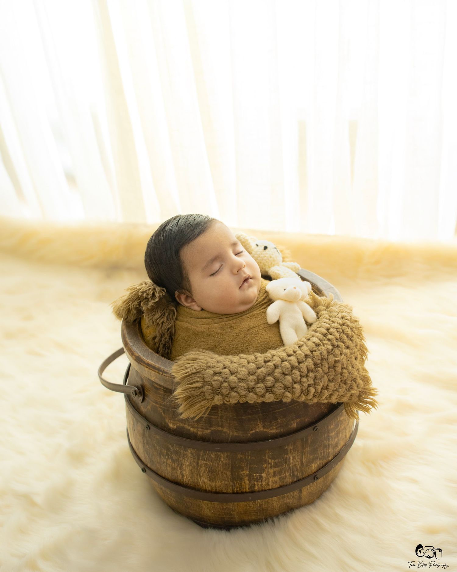 Newborn baby sleeping peacefully in a rustic wooden barrel with a knit blanket and small white teddy bear.
