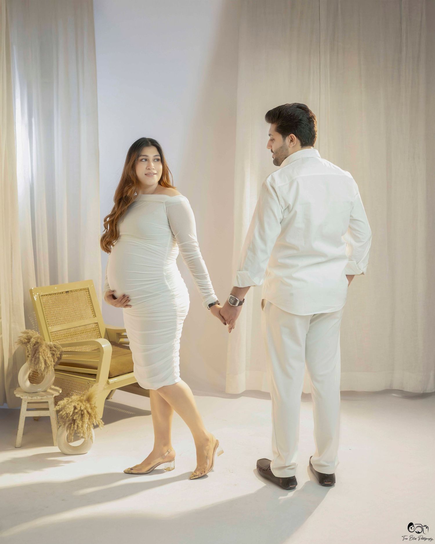 A couple in all-white outfits poses together in a minimalist studio setting with a rattan chair as decor.